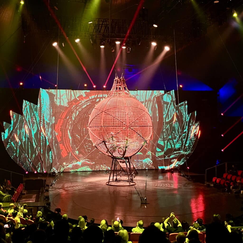 Motorcycle dives around inside of cage on circus stage.